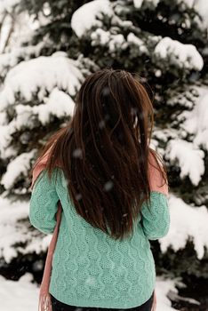 Merry Christmas and happy New Year. Rear view of beautiful brunette woman in pink warm winter clothes standing outdoors in snowy day by christmas tree shaking hair, snow falling