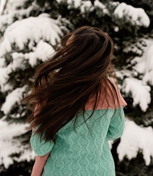 Merry Christmas and happy New Year. Rear view of beautiful brunette woman in pink warm winter clothes standing outdoors in snowy day by christmas tree shaking hair, snow falling