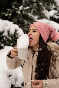 Merry Christmas and happy New Year. Winter fun and joy. woman in warm winter clothes standing by the big christmas tree outdoors and licking snow from the cup