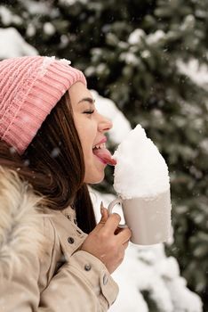 Merry Christmas and happy New Year. Winter fun and joy. woman in warm winter clothes standing by the big christmas tree outdoors and licking snow from the cup