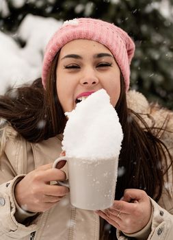 Merry Christmas and happy New Year. Winter fun and joy. woman in warm winter clothes standing by the big christmas tree outdoors and licking snow from the cup