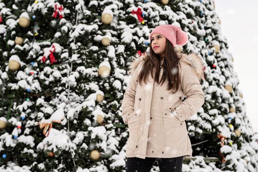 Merry Christmas and happy New Year. Happy Woman in warm winter clothes standing by the big christmas tree outdoors, snow falling