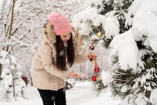 Merry Christmas and happy New Year. Beautiful woman in warm winter clothes decorating Christmas tree in a park in snowy day