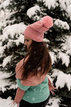 Merry Christmas and happy New Year. Portrait of beautiful brunette woman in pink warm winter clothes standing outdoors in snowy day by christmas tree shaking hair, snow falling
