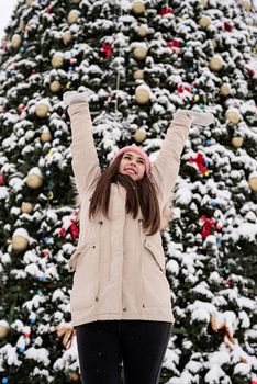 Merry Christmas and happy New Year. Happy Woman in warm winter clothes standing by the big christmas tree outdoors, arms up, snow falling