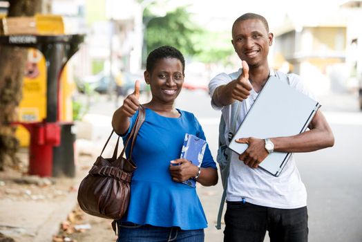 Young happy students with books and computer outdoors. Young couple is showing thumbs up and good luck. Smart young man and a girl in the campus of the University. Apprenticeship and education for young people.