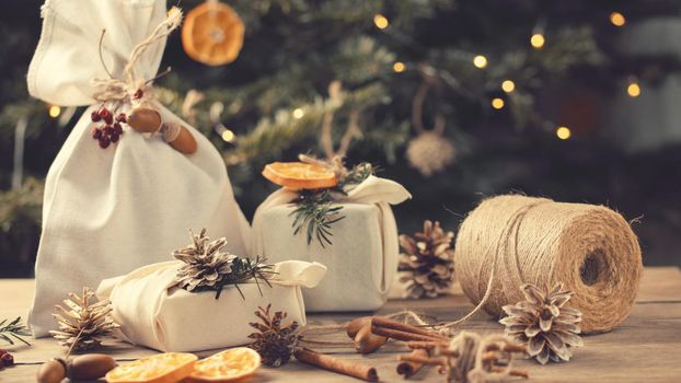 Zero waste christmas concept. Packed in natural fabric gifts and decorations from natural materials on wooden table near Christmas tree with lights.