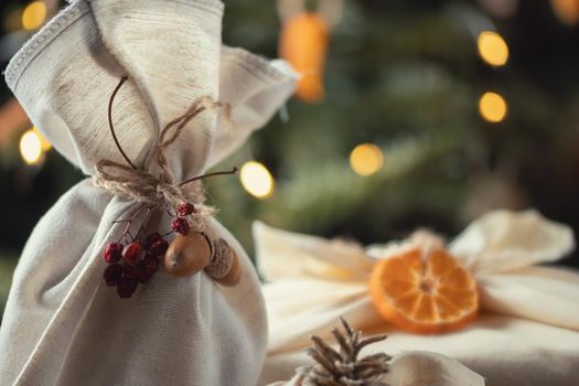 Zero waste christmas concept. Packed in natural fabric gifts and decorations from natural materials on wooden table near Christmas tree with lights.