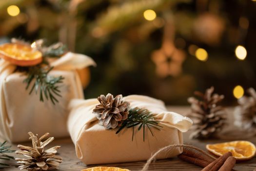 Zero waste christmas concept. Packed in natural fabric gifts and decorations from natural materials on wooden table near Christmas tree with lights.