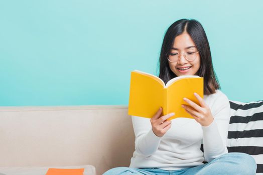 Asian beautiful young woman wearing eyeglasses sitting on sofa reading storybook, portrait relaxation of happy female smiling in living read a book studio shot isolated on blue background