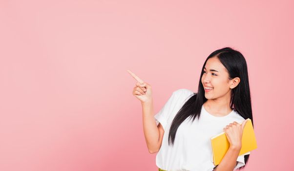 Portrait of happy Asian beautiful young woman confident smiling holding orange book open pointing finger to side copy space, studio shot isolated on pink background, education concept