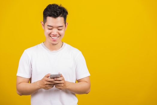 Asian handsome young man smiling positive using smartphone to get in touch screen or typing message, male reading good news message enjoy mobile application, studio shot isolated on yellow background