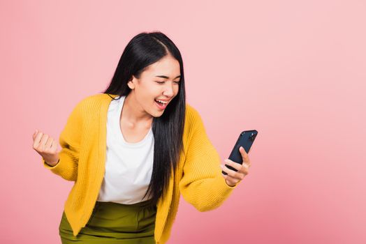Happy Asian portrait beautiful cute young woman teen smiling excited   using mobile phone say yes! Studio shot isolated on pink background, Thai female surprised make winner gesture on smartphone