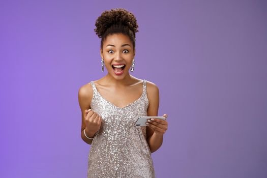 Excited charming young african-american woman lucky winning game playing smartphone standing pleased yelling happily clench fist triumphing joyfully celebrating great news received message.