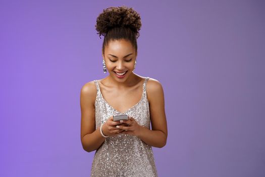 Luxurious good-looking stylish african american woman in silver evening dress holding smartphone chatting texting friends smiling broadly waiting girlfriend near club entrance communicating phone.