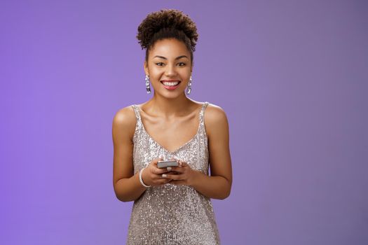 Charming elegant smiling lucky african-american woman in glittering silver dress holding smartphone grinning gladly camera texting using phone send photos after party, standing blue background.