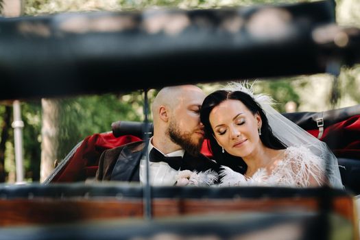 The bride and groom with a bouquet are sitting in a carriage in nature in retro style.