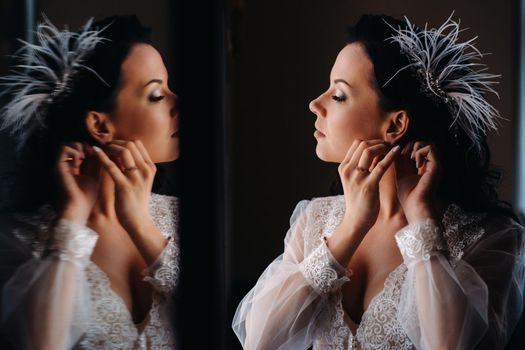 the bride, dressed in a boudoir transparent dress and underwear, wears earrings at the mirror.