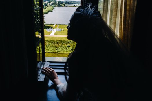 The silhouette of the bride at the window looks at the place of her wedding ceremony on the street.