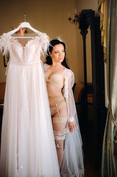 a bride dressed in a boudoir transparent dress and underwear holds her wedding dress in her hands in the interior of the house.