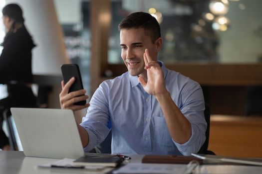 Cheerful happy businessman using smartphone app for virtual communication, holding phone in hand and waving at webcam, holding video call sitting at the desk