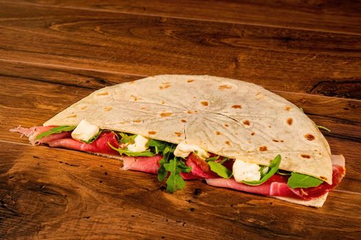 salty piadina with cured meats and vegetables on a wooden surface