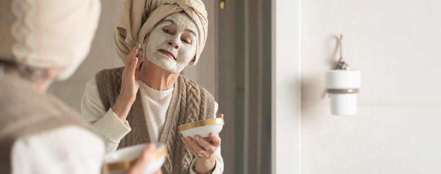 Elderly woman with towel on her head smiles and applies cleansing mask on her face. Happy pensioner looks after her appearance, puts on a white natural mask with collagen, tighten and rejuvenate face.