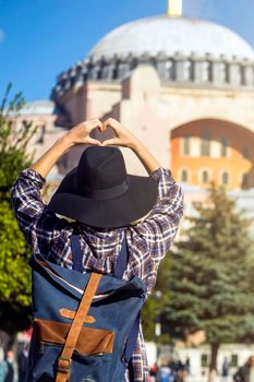 Young traveler in a hat and stylish shirt folds her hands in the shape of a heart against the background of an ancient Hagia Sophia mosque in Istanbul, Turkey. Girl tourist travels to Arab countries.