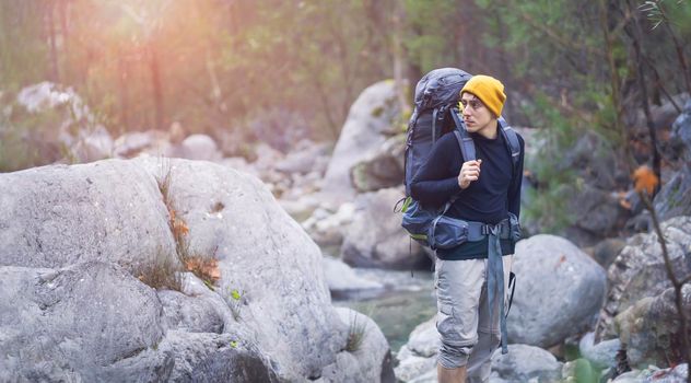 A young smiley man with a backpack and camping equipment stands on the rocks and looks at the beautiful landscape, at the trees and river in a mountain gorge. Trekking the trail in the mountains.