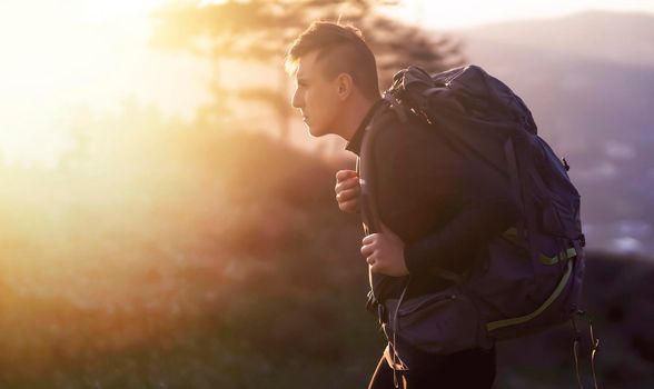Young man with a backpack walks along a trail in the mountains against the backdrop of a beautiful sunset. A traveler is hiking and looking on beautiful view.