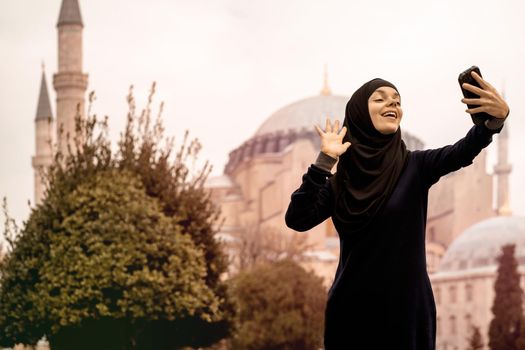 A young smiling Muslim girl in a hijab and stylish black glasses makes a selfie against the background of the Hagia Sophia mosque. Tourism in Istanbul, Turkey