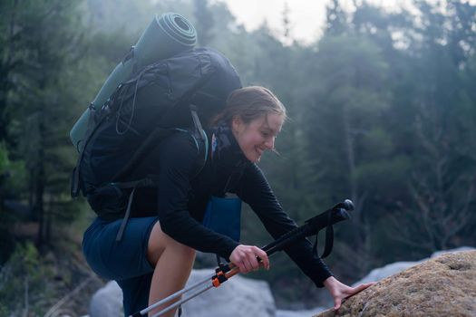 A young smiley girl with a backpack holds trekking poles and climbs a mountain trail in the national park. A hiker explores the wildlife with camping equipment.