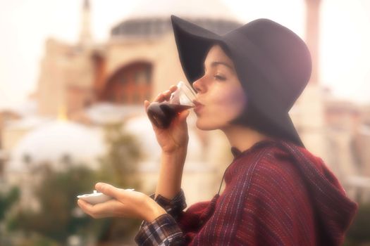 A young girl in a burgundy poncho and a hat holds traditional Turkish tea in hands. Hagia Sophia mosque in the rays of the sunset on the background. Travel in Turkey, Istanbul