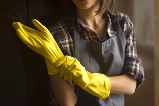 A young girl in a plaid shirt and apron puts yellow rubber gloves on her hands to start cleaning her house and create comfort. Housekeeper with gloves doing disinfection.