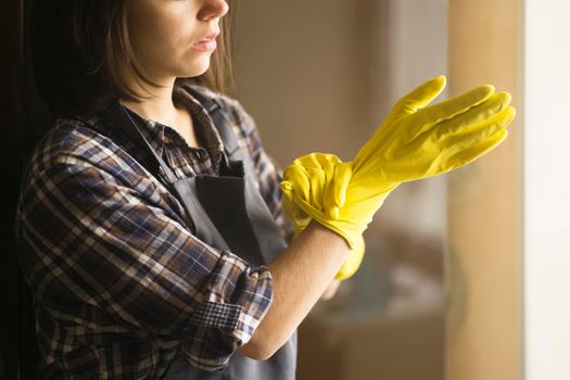 A young girl in a plaid shirt and apron puts yellow rubber gloves on her hands to start cleaning her house and create comfort. Housekeeper with gloves doing disinfection.