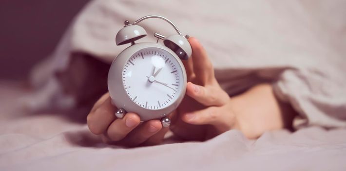 Hands of a young man from under the blankets hold a retro vintage alarm clock in gray. The person holds a clock, need to wake up.