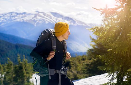 A young smiley man with a backpack and camping equipment stands on the rocks and looks at the beautiful landscape, at the trees and river in a mountain gorge. Trekking the trail in the mountains.