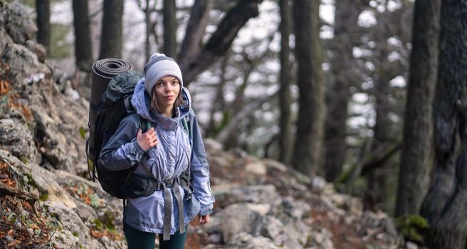 A young girl in a hat and a membrane jacket with a backpack climbs the trail up the mountain in early spring. The traveler is engaged in hiking in the national park.