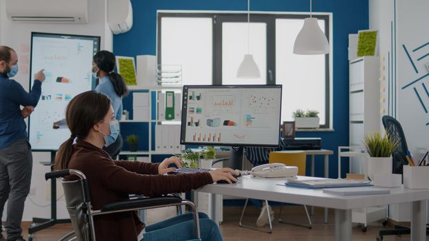 Paralysed financial expert wearing face mask sitting in wheelchair analysing economy statistics of company in new normal business office typing on computer. Handicapped woman using modern technology