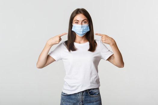 Covid-19, health and social distancing concept. Attractive brunette girl in medical mask pointing finger at face, white background.