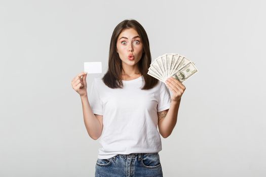 Amused brunette girl looking excited, holding money and credit card, standing white background.