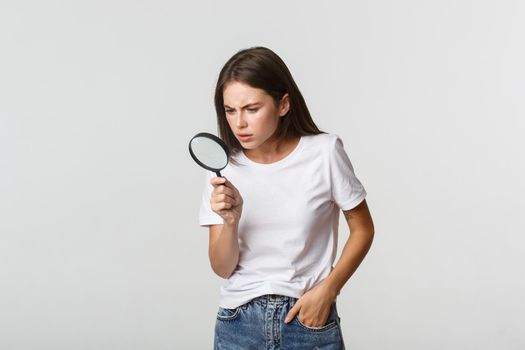 Serious-looking smart beautiful girl looking through magnifying glass down, searching for something.