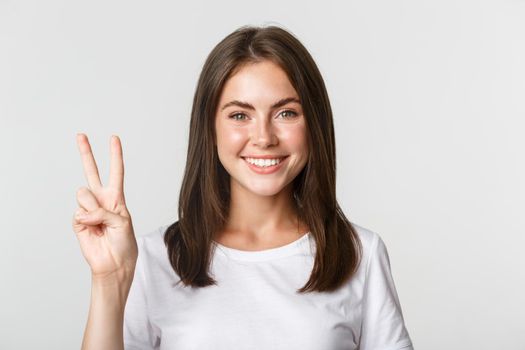 Portrait of beautiful and happy brunette girl smiling and showing peace gesture, standing white background.