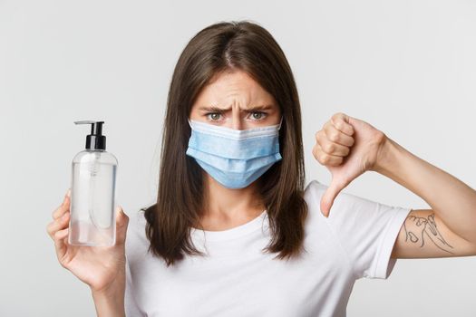Covid-19, health and social distancing concept. Close-up of disappointed cute brunette girl in medical mask, disapprove hand sanitizer, showing thumbs-down.