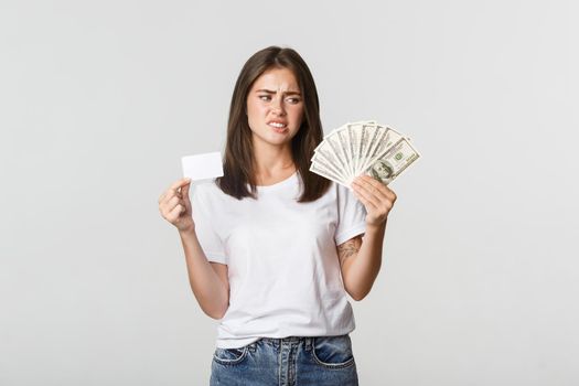 Confused and displeased young woman looking at money and frowning, prefer credit card.