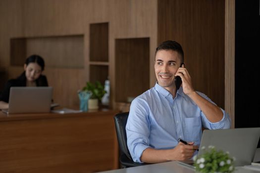 Portrait of young smiling cheerful entrepreneur in modern office making phone call while working with laptop