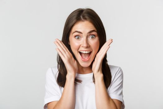 Close-up of excited happy brunette girl looking amazed, holding hands near face astonished.