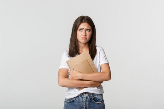 Portrait of sad and disappointed cute female student holding notebooks and looking upper left corner upset.