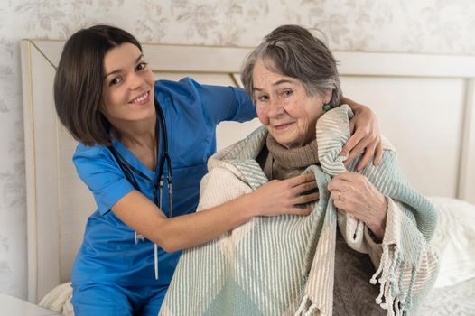 A young nurse takes care of an elderly 80-year-old woman at home, wraps a blanket around her. Happy retired woman and trust between doctor and patient. Medicine and healthcare.