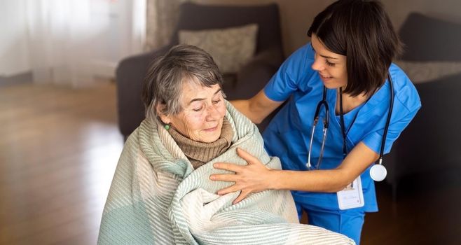 A young nurse takes care of an elderly 80-year-old woman at home, wraps a blanket around her. Happy retired woman and trust between doctor and patient. Medicine and healthcare.